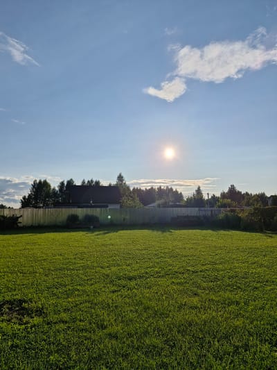 The image depicts a sunlit outdoor scene in the city of Sorochkino, Russia. In the foreground, a lush green lawn stretches across the frame, casting soft shadows from the late afternoon sun. A corrugated metal fence runs horizontally across the middle ground, separating the lawn from a line of trees and a building beyond. The trees are a mix of deciduous and coniferous varieties, with their foliage appearing dense and green. A dark-roofed house is partially visible behind the trees, with a chimney on its roof. The sky above is a clear blue, dotted with a few wispy white clouds, and the sun is a bright, radiant orb in the upper center of the frame, creating a lens flare. The overall impression is one of a peaceful, rural or suburban setting bathed in warm sunlight. There are no people or animals visible in the image.