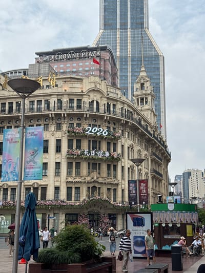 This is a medium-shot, eye-level, outdoor photograph taken on a cloudy day. The scene is a street in Shanghai, China, featuring a blend of historic and modern architecture. In the foreground, a large blue umbrella is closed and leaning against a lamppost. Several people are walking in the street, including a woman holding a red umbrella, two men walking side-by-side, and a man in a wheelchair.
Dominating the center of the image is a grand, ornate building with a light-colored facade, several stories high, featuring balconies, windows with dark shutters, and decorative elements. Above the facade, the "CROWNE PLAZA" hotel sign is visible. In front of this building, banners promoting "SHANGHAI SUMMER 2026" are displayed. A large illuminated sign with the number "2026" is prominently featured on the building's facade, surrounded by pink floral decorations. To the right of this building, a very tall, modern skyscraper with a glass facade rises into the sky. The background shows more modern buildings.
The ground is paved, and there are several planters with greenery. To the right, there's a small kiosk-like structure with bottles on display. The overall atmosphere is bustling with pedestrian activity amidst the cityscape.