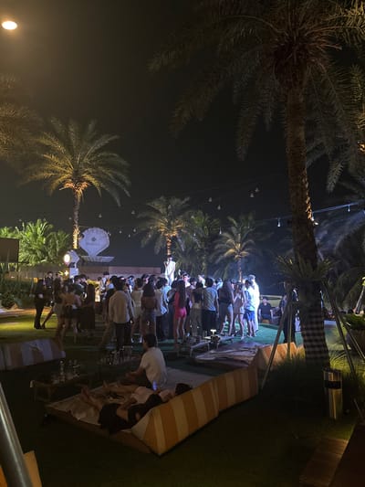 The image shows a nighttime outdoor gathering in Kosambi Barat, Indonesia. Many people are standing closely together, suggesting a social event. Palm trees are visible in the foreground and background, illuminated by scattered string lights and ground lights. Some people are gathered around what appears to be a bar area, with glassware visible on the surface. The lighting conditions are low, creating a soft glow around the gathering. There are covered sitting areas, and some individuals are seated in them.