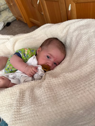A baby is lying on a soft, cream-colored, textured blanket. The baby is on its stomach, facing left, with its head turned towards the camera. It has dark hair and blue eyes and is holding a brown pacifier in its mouth with its left hand. The baby is wearing a green shirt with a pattern of cars and has a white cloth under its head. In the background, a wooden dresser with two drawers and silver handles is visible. The location is identified as Vylygort, Russia.