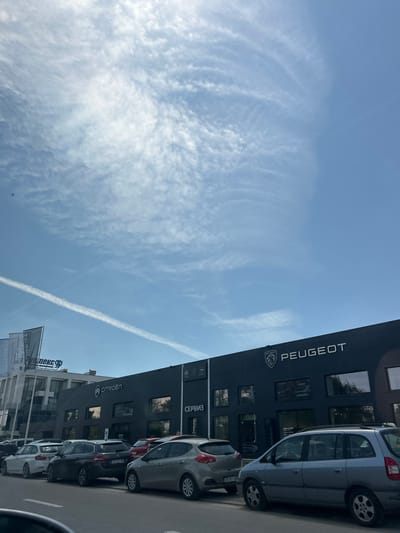 The image depicts a car dealership in Lozen, Bulgaria, featuring the brands Peugeot and Citroën. The building is modern, with dark grey facades and large windows. The Peugeot logo and name are prominently displayed across the top of the building. Next to it, the Citroën logo and name are visible. The dealership appears to be a service center as well, indicated by the word "СЕРВИЗ" (service in Bulgarian) on the building. Several cars are parked outside, including what appear to be newer models, suggesting a functioning dealership. The sky is blue with scattered white clouds and a contrail from an airplane. The overall scene suggests a bright, sunny day.