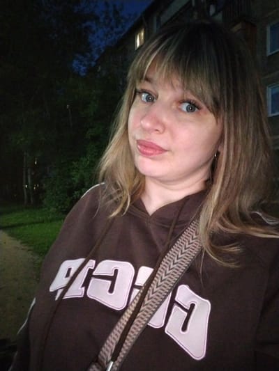 A woman is taking a selfie outdoors at dusk. She has blonde hair with bangs and blue eyes, and is wearing a brown hoodie with white lettering. A strap from a bag is visible across her chest. Behind her, a dark tree line is visible, with a building with lit windows in the background. The scene appears to be a residential area. No specific location in Yaroslavl, Russia can be identified from the image alone.