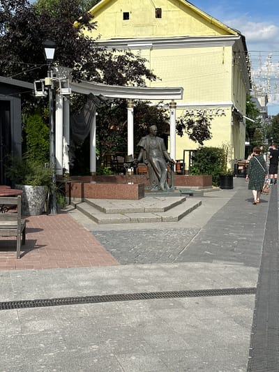 A full shot captures a sunny day in what appears to be a public square or pedestrian area in Putyatino (Semivragovskaya S/A), Russia. A statue of a man in academic attire, leaning on a pedestal, is a central focus. Behind the statue, a light yellow building with a decorative cornice stands, featuring a sign with Cyrillic lettering. The ground is paved with cobblestones and granite slabs, with a prominent grate running across the foreground. To the right, two women walk away from the camera, one carrying a shopping bag. To the left, outdoor seating and planters are visible, suggesting a cafe or restaurant setting. Lush green trees and a lamppost add to the urban landscape.