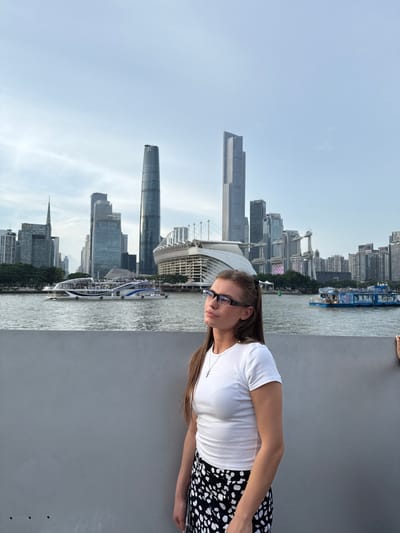 A young woman with long brown hair, wearing glasses, a white t-shirt, and a black and white patterned skirt, stands in the foreground looking upwards and to the right. She is positioned in front of a grey wall. Behind her, a wide river flows, with several boats on its surface. In the background, a modern cityscape unfolds, featuring numerous skyscrapers of varying heights and architectural styles, along with a large stadium-like structure. The sky is overcast. The location appears to be the Pearl River waterfront in Yuexiu District, China, likely in the city of Guangzhou, given the prominent skyscrapers like the Canton Tower and the Guangzhou IFC.