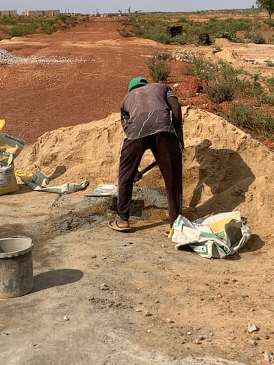 A man wearing a green head covering, a grey t-shirt with dark patterns, and dark pants is standing outside at a construction site in Kujama, Nigeria. He is near a pile of dirt/earth/sand, some open cement bags, and a piece of construction equipment. The background shows buildings and vegetation.