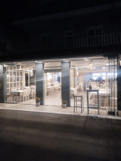 The image depicts the exterior of a restaurant at night in Neoi Epivates, Greece. The restaurant features a glass-enclosed dining area with tables and chairs. The interior is illuminated, revealing white tablecloths and chairs, suggesting it is prepared for or currently serving patrons. There is decorative fencing or screens on the left side of the interior. Two potted plants, one with orange flowers and another with a single green plant, are visible outside the entrance. A floor fan is positioned inside the restaurant. The street in front of the restaurant is dark and paved. The building above the restaurant appears to be residential, with balconies and windows visible.