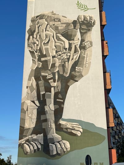 This media file depicts a large mural painted on the side of a building. The mural is a monochromatic, cubist-style artwork featuring a stylized, robotic-looking figure, possibly a giant or a cyborg. The figure is depicted in a dynamic pose, with its arms outstretched and fists clenched. The mural is rendered in shades of gray and beige, with subtle shading to create a sense of depth and texture.

Scattered throughout the mural are various words in raised, blocky lettering. Some of these words are legible and appear to be place names, such as "LA MANCHE," "NARVIK," "MURMANSK," "OSTSIE," "CHOM TERCH," and "KOST ZEZL." These names suggest a connection to historical events or geographical locations, possibly related to war or exploration. A small green leaf sprig is painted to the left of the figure's head, contrasting with the stark, industrial aesthetic of the main artwork.

The mural is set against a bright blue sky, indicating it is daytime. The sunlight casts shadows on the mural, highlighting its three-dimensional qualities. The top of the image shows the roofline of buildings behind the mural, including angled roofs and some industrial elements, suggesting an urban environment. The overall mood of the mural is powerful and perhaps even imposing, with the monumental scale and geometric forms. The presence of the leaf might suggest a theme of nature reclaiming or coexisting with industrial might. The lettering "MARX" is visible vertically on the right side of the mural.