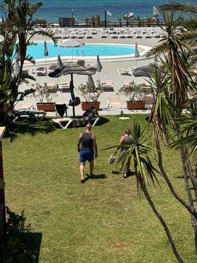 The image depicts an outdoor scene at a resort with a swimming pool and a grassy area. In the foreground, two individuals are walking on the lawn. A man wearing a black tank top and blue shorts is walking away from the camera. A woman in a grey outfit, carrying a bag, is walking in the same direction. The background shows a swimming pool with lounge chairs and umbrellas surrounding it, and beyond that, the sea. The environment is sunny and appears to be a vacation setting, likely a hotel or resort in Isola delle Femmine, Italy.