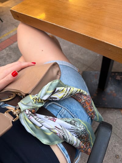A person, identified by their legs and hand with red nail polish, is seated at a wooden table outdoors, likely in İzmir, Turkey. The individual is wearing denim shorts over a dark garment and holding a light brown clutch purse adorned with a colorful scarf tied to its strap. The purse has a repeating embossed logo pattern and a zipper closure. The scarf features an abstract print with botanical and fruit motifs. A dark metal chair leg is visible to the right, and the table has a light brown, wooden surface. The background shows a textured sidewalk and a brick border.