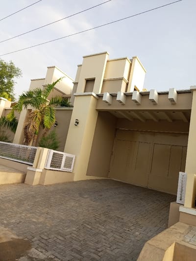 The image shows a large, beige-colored, modern house with a carport in Maiduguri, Nigeria (latitude: 11.82, longitude: 13.154). The house has a white awning above the carport entrance, and a large, double-door garage door. The garage door is closed and made of tan-colored metal. There is a palm tree in front of the house, and a small, white ventilation grate on the side of the house. The yard is covered in gray cobblestones. The sky is a light blue and there are power lines running above the house. There are no people visible in the image.