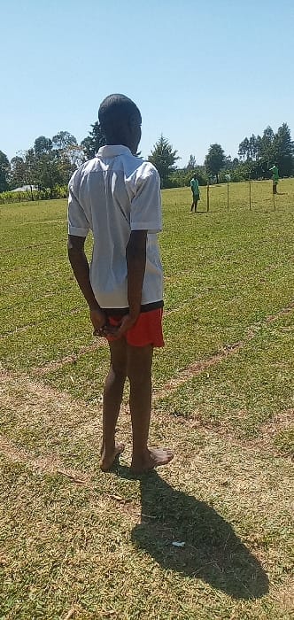 The image shows a young boy standing on a grassy field in Burundu, Kenya, at coordinates 0.53 latitude and 34.746 longitude. The boy is wearing a white shirt and red shorts. He has his hands clasped behind him, and he is looking at two other people in the distance. The field is covered in short green grass. Two other figures are visible in the distance, moving away from the boy. The sky is clear and blue. The image appears to be taken on a sunny day. The trees in the background are mostly deciduous. The field is relatively flat and without noticeable obstructions. The scene is peaceful and serene.
