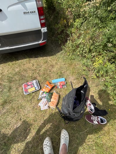 A black backpack lies open on grass, surrounded by items including two water bottles, a bottle of soda, a loaf of bread, snacks like chips and sausage, and a sealed container. Nearby, a pair of maroon sneakers with black socks sit on the grass. The back of a white van is partially visible on the left. Two feet wearing white Crocs are situated on the right, adding a human presence to the scene.