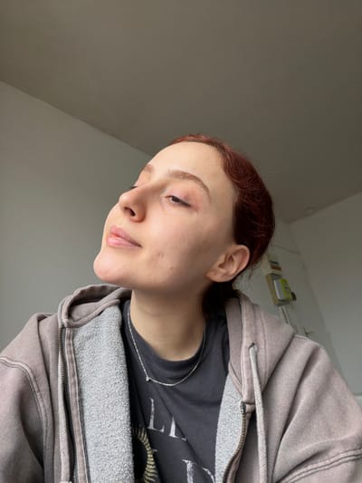 The image depicts a young adult female in an indoor setting within Reims, France. The subject, with reddish-brown hair pulled back, is positioned with her head tilted upward and to her right, eyes partially closed and gaze directed slightly left and upward. She is wearing a dark grey t-shirt with a faint graphic design visible on the chest, layered under an unzipped, light brown or taupe hooded sweatshirt. A thin, silver-colored chain necklace with a small, circular pendant is visible around her neck. The background consists of a plain, light grey wall. A slight shadow or darker tone is present in the upper right portion of the background, and a small segment of a light-colored, wall-mounted object or shelf with indistinct items is visible on the far right edge of the frame.