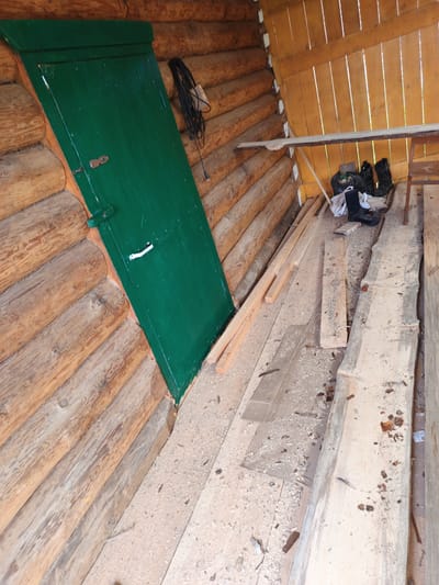 The image depicts the interior of a rustic structure, likely a shed or storage area, constructed from rough-hewn logs. A prominent, emerald-green door is visible on the left, angled with the slope of the building. The door has a simple latch and a white handle. To the right of the door, a coiled black cable with a white electrical plug hangs from the wall.
The floor of the structure is made of wooden planks, covered in sawdust and wood shavings, suggesting recent carpentry or construction work. Various wooden planks, some rough-cut and others appearing to be reclaimed lumber, are scattered across the floor.
In the background, a wooden shelf made of a single plank is supported by metal legs. Underneath this shelf, a black plastic bag filled with debris is present, along with a pair of black rubber boots and another boot lying on its side. To the far right, a simple wooden chair with a metal frame is visible, partially obscured by a wooden plank. The walls and ceiling are also constructed from wooden planks, with a warm, yellowish hue. The overall impression is one of a working space or storage area that is in a state of disarray. The location is identified as Islambaevo, Russia.