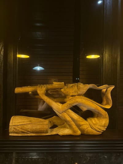 A wooden sculpture is displayed indoors in Tibubiyu, Indonesia. The sculpture, crafted from light-colored wood, depicts a stylized human figure in a semi-reclined position, actively playing a wind instrument held to its mouth with both hands. An additional cylindrical wooden object, resembling a drum, rests on the display surface to the left of the figure. The entire arrangement is situated on a dark, reflective base, likely marble, and is positioned behind a glass panel. This panel reflects overhead light sources, specifically a distinct yellow, oval-shaped light in the upper left and a white, semi-circular light lower and more centrally located. The background consists of a dark, horizontally paneled wooden wall.