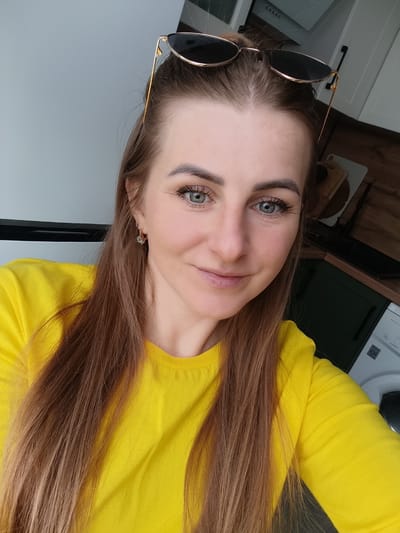A woman is taking a selfie indoors. She has long brown hair, green eyes, and is wearing a yellow t-shirt. Sunglasses are resting on top of her head. The background includes a white wall, kitchen cabinets (some dark green), and a washing machine. The location is in a residential space in Chernogolovka, Russia.