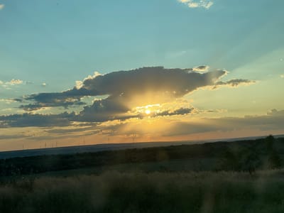The image depicts a sunset over a rural landscape, likely in or near Colibași, Moldova. The sky is a gradient of blues and yellows, with clouds illuminated by the setting sun. Sunbeams radiate from behind a large, dark cloud, creating a dramatic effect. In the distance, a row of wind turbines stands silhouetted against the horizon. The foreground is dominated by a field of tall, dry grass. The overall scene is serene and natural, with no people or specific man-made structures immediately visible apart from the wind turbines.