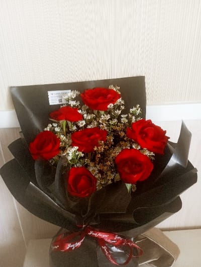 A bouquet of seven red roses nestled among smaller white flowers is wrapped in elegant black paper. A red ribbon with the inscription "Just for you" is tied around the bouquet, adding a touch of sophistication. The bouquet leans slightly against a light-colored wall, creating a simple yet striking composition. The dark wrapping contrasts with the vibrant red of the roses, highlighting their beauty.