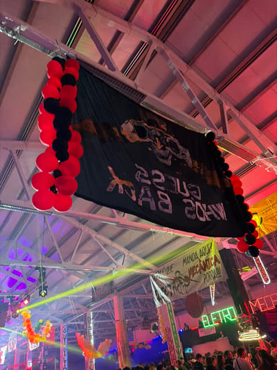 This image captures an indoor nighttime event, likely a university or student gathering, held within a large hall featuring an industrial-style ceiling with prominent metal trusses and beams. The space is vibrantly decorated with numerous banners, balloons, and colorful lighting.
A large black banner hangs centrally from the ceiling, adorned with clusters of red and black balloons at its top and bottom. This banner displays a graphic that appears to be a stylized skull or head, along with inverted white text, partially legible as "MADE BY 2024".
Around the venue, several other banners are visible, suggesting different student groups or academic departments. A yellow banner reads "NÚCLEO D DE DESPO INSTITUTO DE BRAGH," directly referencing the Instituto Politécnico de Bragança. Another light green banner proclaims "QUEM É QUE MANDA AQUI? MECÂNICA PORRA!!!" with a gear-like logo, indicating a mechanical engineering student association. Further in the background, banners with "CONTABILID" (likely Accounting) and "LINEAB" are discernible.
The atmosphere is festive, illuminated by dynamic lighting, including red, purple, yellow, and green spotlights casting beams across the space. Neon signs spell out "ELETRO" and "ELETROMED". On the far right, a crowd of people can be seen, possibly observing the event from an elevated vantage point, underscoring the social and celebratory nature of the occasion. Red, black, yellow, orange, and white balloons are arranged throughout, with some forming an arc shape in the background. The event appears to be a lively student party or festival in Bragança, Portugal.