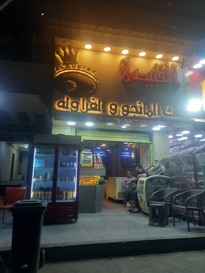 The image captures a street view at night in Cairo, Egypt. A brightly lit storefront dominates the left side of the frame. The signage features Arabic script, illuminated with warm lights, and a logo. Below the signage, there is the name "Alqubisy." A refrigeration unit filled with beverages and drinks is in the foreground. Further inside the store, there are fruits and juice dispensers. Two men are seated near the store entrance, one of them wearing a purple shirt. Several chairs are stacked on the right side. The overall lighting creates a sharp contrast between the illuminated storefront and the darker surroundings.
