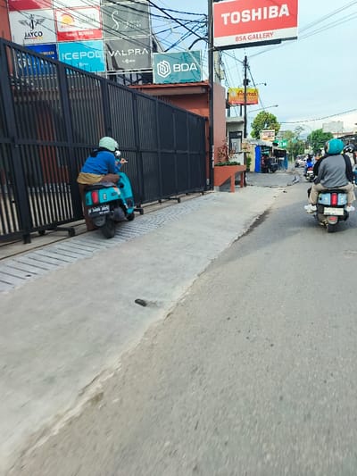 The scene is in Katapang, Indonesia, at coordinates -6.989, 107.558. Two motorcyclists are on the road, both wearing helmets and driving in the same direction. One motorcyclist is parked near a closed black gate, while the second motorcyclist is driving past. The street is lined with businesses, with multiple signs visible, including one for "TOSHIBA BSA GALLERY." The road is paved and has a slight incline. A small sidewalk is present along the side of the road, with some greenery and a concrete curb. The sky is clear with a few visible power lines overhead. The scene appears to be a quiet, normal street scene. There is no one else visible in the image.