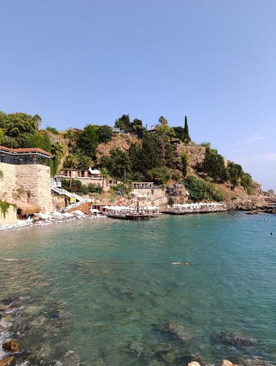 This image depicts a scenic coastal view in Antalya, Turkey, specifically likely from Mermerli Beach. The foreground shows clear, turquoise water with visible rocks beneath the surface. Several people are swimming and enjoying the water. A rope with buoys stretches across a section of the water.
Along the shoreline, a sandy beach is lined with numerous white lounge chairs and umbrellas, indicating a resort or public beach area. A wooden pier extends over the water, also furnished with seating and umbrellas. Buildings are nestled into the rocky hillside that rises behind the beach. These structures appear to be part of a restaurant or complex, with one prominent building featuring a stone base and a red-tiled roof. White staircases and pathways connect the different levels of the hillside.
The hillside itself is covered in lush green vegetation, including trees and shrubs, adding to the natural beauty of the location. The sky is a clear, bright blue, suggesting a sunny day. The overall impression is a vibrant and relaxing seaside environment in a Mediterranean setting.