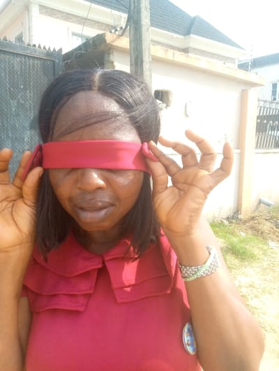 A woman with dark hair has a red cloth tied around her eyes, acting as a blindfold. She wears a red collared shirt and a silver and green watch on her left wrist. She's standing outside, with a building and fence visible in the background. She holds the ends of the blindfold with her hands, appearing as if she is either tying it or about to remove it.