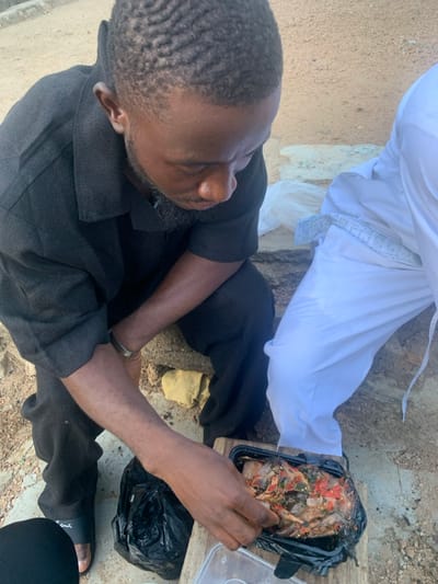 In Okene, Nigeria, an outdoor scene shows two individuals on unpaved ground with concrete elements. A Black male, with a short, wavy haircut and a beard, wearing a dark short-sleeved collared shirt, dark pants, and dark slide sandals, is squatting. He wears a silver-colored bracelet on his left wrist. His right hand is reaching into a black rectangular plastic container holding a prepared meal of fish or seafood stew with visible red peppers and green herbs. Adjacent to this, a clear plastic container lid is on the ground. A black plastic bag is positioned near the food container. To the right, partial view of a second person shows legs clad in white pants and a white garment with a light-colored patterned trim. A white plastic bag and a yellow cloth are visible on the sandy ground in the background. The primary male subject is looking down at the food container.