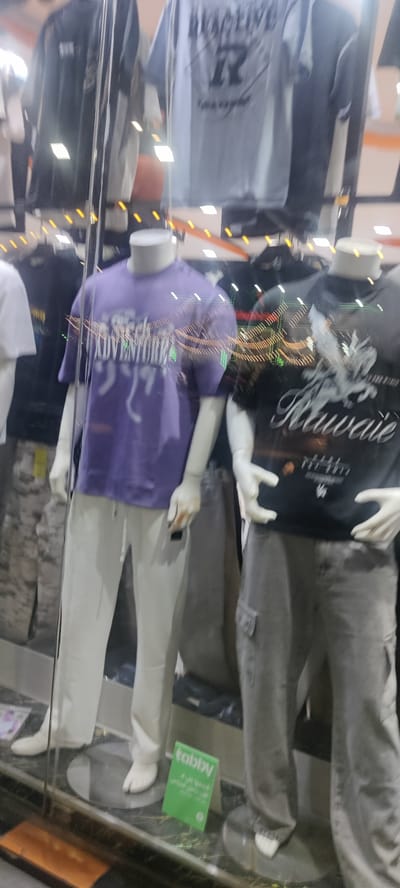 The image shows a storefront display of mannequins in Jeddah, Saudi Arabia. Two mannequins are prominently featured. The one on the left wears a purple t-shirt with a graphic design, paired with white sweatpants. The mannequin on the right is dressed in a black t-shirt with a design featuring the word "Kawaie", and gray cargo pants. Other clothing items are visible in the background, displayed on hangers. A green sign with the word "tabby" is placed in front of the mannequin on the left. The scene is viewed through a glass storefront, with light reflections visible on the glass. The surrounding environment is a retail setting, likely a clothing store in Jeddah.