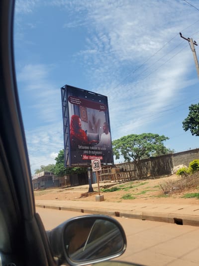 The image depicts a roadside scene in Television, Nigeria, captured from inside a vehicle, with portions of the car's dark interior and side mirror visible in the foreground. The sky is bright blue with scattered white clouds.
A large, double-sided digital billboard stands prominently on the left side of the road. The visible side displays an advertisement featuring two individuals: a woman in a red hijab and patterned top, seated on a light-colored sofa while looking at an unseen device; and a man in a white t-shirt, also seated on a sofa, looking towards the woman. The advertisement contains text in Hausa, which translates to: "I want us to discuss tomorrow's worries/concerns" (top left), "Discussion between spouses is important" (bottom center), and "Let's discuss" (bottom center). The MTN logo is partially visible in the top right corner of the billboard.
Adjacent to the billboard, a circular speed limit sign indicates "30 KM/H". The immediate road surface appears to be packed reddish-brown dirt, transitioning to a paved asphalt road beyond. A concrete sidewalk with multiple rectangular drainage openings runs along the right side of the dirt road.
In the background on the left, several low-rise commercial structures with corrugated metal roofs are visible, one sign partially displaying "SAVEX". On the right side, beyond the road and sidewalk, a concrete wall topped with barbed wire runs horizontally, backed by various trees and sparse green vegetation. A concrete utility pole with power lines extends into the sky on the far right. The overall lighting suggests bright daylight conditions.