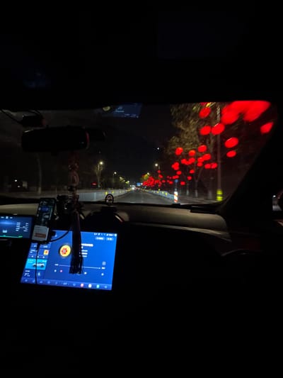 This media file captures a nighttime scene from the interior of a car, looking out through the windshield and towards the passenger side. The primary subject is a street adorned with numerous bright red, circular lights, likely lanterns or festive decorations, strung along trees or poles, creating a vibrant, illuminated pathway. These lights are slightly out of focus, indicating they are outside the vehicle. A road is visible with white lane markings and what appears to be a yellow barrier or curb line.

Inside the car, a large, upright digital screen with a blue background is prominently displayed on the right, showing a user interface with various icons and text, which appears to be in Chinese. Below this main screen, a smaller electronic device, possibly a smartphone, is mounted and visible. A microphone on a flexible arm extends towards the center of the car, suggesting use for voice commands, calls, or recording. Cables connect these devices. A decorative item, resembling a string of red and clear beads or perhaps a food skewer, hangs near the dashboard.

The time of day is clearly night, with the surroundings outside the illuminated street appearing very dark. No people are visible in the image. The scene suggests driving through a decorated area in Shenhe District, China, possibly during a holiday or festival, with the car's interior configured for navigation, entertainment, or communication.