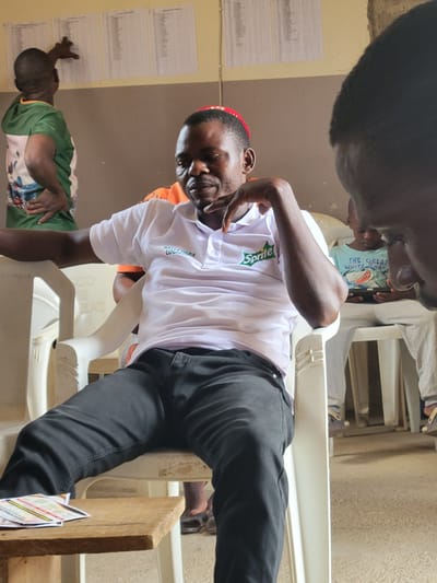 A man in a white Sprite-branded shirt and black pants sits in a white plastic chair in an indoor setting, possibly a room in Madalla, Nigeria. He is making a hand gesture with his right hand. The background includes walls with papers attached, and other people are present, sitting in chairs. The scene appears to be a casual meeting or gathering.
