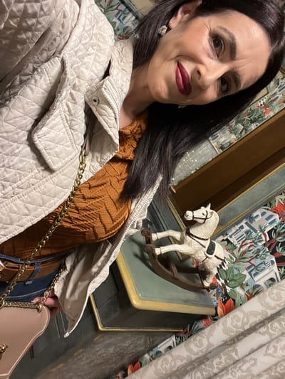 The image depicts a woman with dark hair, wearing dark lipstick, posed for a photo. She appears to be wearing a beige quilted jacket and a brown patterned top. A handbag with a gold chain is visible. A decorative rocking horse is positioned near a piece of furniture. The background features wallpaper with a floral or ornate pattern. The location might be inferred from the prompt as Konz, Germany.