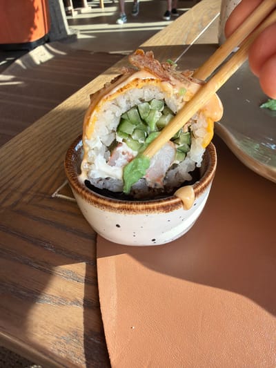 In Khotianivka, Ukraine, a close-up shot captures a hand using chopsticks to lift a piece of sushi from a small, speckled ceramic bowl. The sushi roll is topped with a creamy orange sauce and crispy bits, and its interior reveals rice, cucumber, and what appears to be crab or imitation crab meat. A dollop of bright green wasabi is also visible. The sushi rests on a wooden table, with a portion of a brown textured placemat in the foreground. Sunlight casts distinct shadows across the table, suggesting an outdoor or well-lit indoor setting. In the blurred background, the legs of people in casual attire are visible, and part of an orange cylindrical object, possibly furniture, is present.