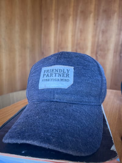 The image depicts a dark blue fabric baseball cap resting on a dark object, which is situated on a light brown wooden surface. The cap features a light-colored rectangular patch on its front, displaying the text "FRIENDLY PARTNER" above "CORE YOUR MIND." The background consists of a vertical light brown wooden wall with visible grain. A partially visible, out-of-focus dark object is present to the right of the cap. The scene is located in the city of Gyellesu, Nigeria.