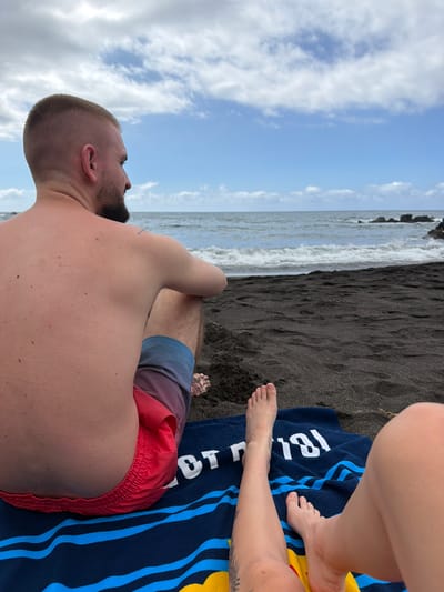 In Puerto de la Cruz, Spain, a man with a buzz cut and beard sits on a black sand beach, facing the ocean. He wears red and blue swim trunks. Beside him, on a blue towel with yellow and blue stripes, rest a woman's bare legs and feet. The ocean is visible in the background, with waves gently rolling towards the shore. The sky is partly cloudy with a mix of blue and white.