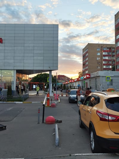 In Ramenskoye, Russia, a street scene unfolds under a dramatic sky with illuminated clouds. The foreground is dominated by the rear quarter of a yellow taxi, its license plate obscured. To the left, a dark-colored car is parked, and two men are standing next to it, seemingly engaged in conversation.

Further down the street, a modern building with a silver facade occupies the left side of the frame, its glass entrance revealing interior lights and a few individuals. Beyond this, a row of smaller shops with illuminated signs stretches into the distance. Several people are visible near these shops, and a yellow traffic sign indicating a pedestrian crossing is prominent.

The background features a multi-story brick apartment building with red trim around the windows, characteristic of Soviet-era architecture. The overall environment is urban, with paved roads, sidewalks, and various street furniture like bollards and barriers. The time of day appears to be late afternoon or early evening, given the warm hues in the sky.