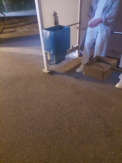 The image shows an outdoor scene in Altdorf, Germany. A person in light-colored clothing is standing next to a metal trash can, near an ashtray filled with cigarette butts. Beside the trash can is a blue plastic bag, likely for waste disposal, and a bicycle is visible in the lower left corner. The ground is paved.