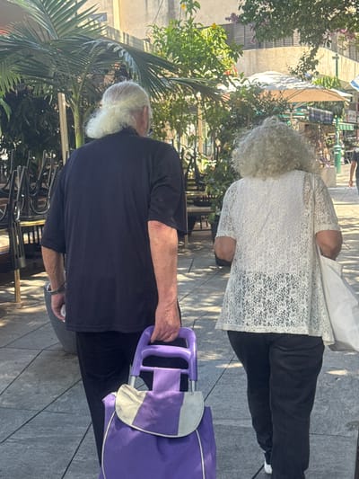 The image shows a man and a woman walking on a paved sidewalk in Ramat Gan, Israel. The man, from the back, has long, light-colored hair pulled back and is wearing a dark t-shirt and dark pants. He is holding the handle of a purple shopping trolley. The woman, also from the back, has curly, light-colored hair and is wearing a white lace top and dark pants. She is carrying a white tote bag over her right shoulder. In the background, there are tables and chairs stacked near a building, lush green plants, and a pale beige umbrella providing shade over an outdoor seating area. Sunlight illuminates the scene, casting shadows on the sidewalk.