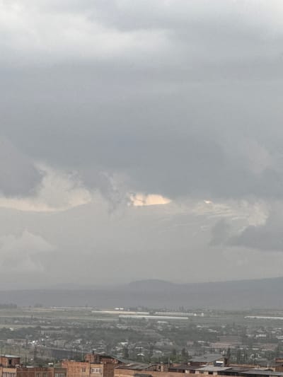 This is a landscape photograph taken from an elevated perspective, possibly from a balcony or a hilltop. The image is oriented vertically, with the right side of the frame showing a more detailed view of a town or city, while the left side is dominated by a cloudy sky.

The town appears to be a mixture of residential and industrial buildings. Several structures with visible windows and roofs are discernible, suggesting a developed urban or semi-urban area. Some parts of the town seem to be lined with trees or greenery. The rightmost edge of the image reveals the facades of some brick buildings, one of which has multiple windows with white frames.

The sky is heavily overcast with dense grey clouds. There is a patch of brighter light breaking through the clouds in the upper central portion of the sky, suggesting either a break in the clouds or sunlight filtering through. The overall lighting is diffused, indicating a cloudy or possibly rainy day.

The scene depicts a town nestled in a valley or plain, with hills or mountains visible in the distance on the left side, partially obscured by the clouds. The weather appears to be overcast and potentially damp. There is no discernible activity or specific event taking place, and no people are clearly visible. The photograph conveys a sense of a quiet, possibly cool, day with limited visibility due to the cloud cover. No text is visible in the image. The location context suggests this is a view of Abovyan, Armenia.