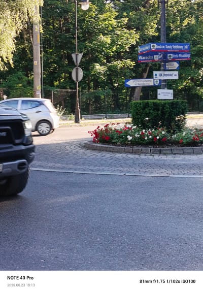 This is a media file captured outdoors in what appears to be a park or urban setting. The scene includes a street signpost with multiple directional signs in Polish, indicating "Warecka," "Ronda im. Danuty Sedzikówny Inki," and "Józefa Pilsudskiego." There are also signs for "SPZZOZ-Przychodnia ul. Warecka 15a" and "STOCER."
In the foreground, the front of a dark-colored vehicle, possibly an SUV, is visible, along with a paved road surface. A white car is seen driving away on the right side of the image. The background is dominated by lush green trees, suggesting a pleasant, possibly sunny day. A circular flower bed with red and white flowers adds a touch of color to the scene. The lighting suggests it is daytime, with sunlight filtering through the trees.
At the bottom of the image, text indicates "NOTE 40 Pro" and the date "2026.06.23 18:13," along with camera settings "81mm f/1.75 1/102s ISO100." The location context provided is Konstancin-Jeziorna, Poland. The overall impression is a snapshot of a street corner or intersection in a Polish town, featuring urban signage, vehicles, and natural elements.