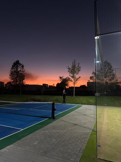 A tennis court sits adjacent to a glass-enclosed area, reflecting the twilight sky. A person stands near the net on the court, surrounded by green grass. The sunset casts a vivid orange and purple hue across the sky. Trees stand silhouetted against the colorful backdrop. The court is neatly bordered with gray brick.
