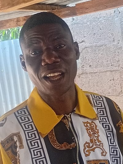 A frontal portrait shot depicts an adult male in Ayangba, Nigeria. The subject has short, dark hair and dark eyes, looking directly at the camera. His mouth is slightly open, revealing several teeth. He wears a collared, long-sleeved shirt with a central metallic zipper. The shirt features a solid yellow collar and upper shoulder sections. The main body of the shirt displays a pattern of alternating vertical panels: white panels with black Greek key borders, and black panels containing intricate gold and brown baroque-style scrollwork and a distinct gold chain motif. The background consists of a light-colored, textured concrete or brick wall on the right, a corrugated metal roof supported by visible wooden beams overhead, and a hint of green foliage on the far left. The subject is stationary.