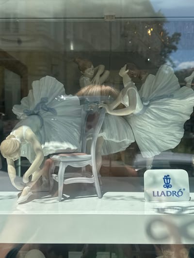 This media file features a close-up, vertically oriented view of three Lladro porcelain figurines of ballerinas, displayed behind glass. The ballerinas are depicted in various graceful poses, adorned in white tutus and bodices. One figurine is prominently in the foreground, while two others are partially visible behind it. The figurines are delicate and detailed, with flowing fabric and expressive postures.

A white Lladro brand plaque with a blue logo and text is visible on the right side of the display, reading "LLADRÓ". The background is blurred and reflects an outdoor scene, suggesting the display is in a shop window or a similar retail environment. The reflections include trees and what appear to be streetlights or interior lighting.

The overall impression is one of elegance and artistry. The lighting creates highlights and shadows on the porcelain, emphasizing the textures and forms of the figurines. The setting appears to be a well-lit, possibly indoor, display area, with the outdoor reflections indicating the location is likely a street-facing window in Karlovy Vary, Czechia. No specific time of day or weather conditions can be definitively determined beyond the reflection of an outdoor environment. The scene evokes a sense of quiet beauty and admiration for the craftsmanship of the Lladro pieces.