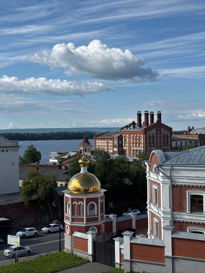 This image captures a street scene in Novokuybyshevsk, Russia, under a bright, partly cloudy sky. The perspective is from an elevated vantage point, looking down at a street lined with buildings and trees. The buildings feature red brick facades with white decorative elements and metal roofs. One prominent building has a golden dome, characteristic of Russian Orthodox churches. Several white cars are parked along the street. The presence of trees suggests it's likely spring, summer, or early autumn. The overall mood is tranquil and urban. The sky is a mix of blue with streaks of cirrus clouds and larger cumulus formations. The time of day appears to be daytime, given the brightness and shadows. One signpost visible on the right has Russian text, but it is illegible due to its size and distance.