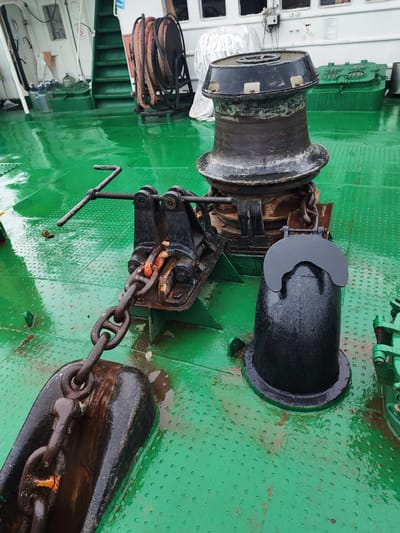 The image shows the deck of a vessel in Vytegra, Russia, with a focus on anchor equipment. A large, dark-colored anchor lies in the foreground, connected by a heavy chain to a black metal mechanism with a lever. Behind this, a substantial black capstan with a grooved drum is visible. To the right of the capstan, a black, bell-shaped object with a curved cover sits on the deck. The deck itself is painted a bright, glossy green and appears wet, with visible water droplets and some reddish-brown staining. In the background, there are green stairs leading up to a white structure, with coiled rope on a reel to the left of the stairs, and a green metal hatch to the right of the capstan. Several large plastic water jugs are also visible near the stairs. The scene suggests a working vessel, likely docked or anchored.