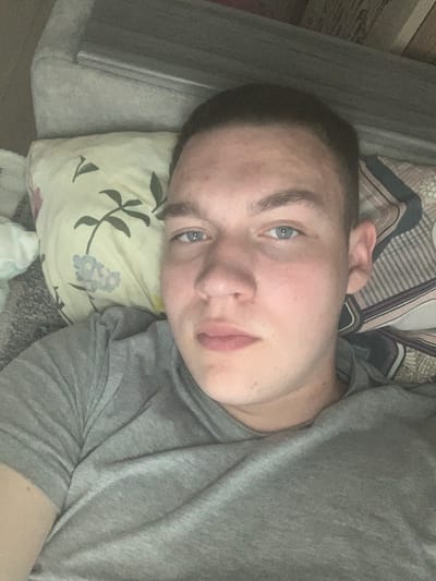 The image is a close-up, horizontally oriented selfie of a young person with fair skin, blue eyes, and light brown hair. They are lying down, resting their head on a pillow with a floral pattern in shades of yellow, green, and light blue. The person is wearing a gray t-shirt. The background includes a gray surface, possibly a couch or bed, and patterned bedding. The lighting suggests an indoor setting, possibly during the daytime, with no distinct indicators of weather or a specific time of day. The person's expression is neutral or slightly relaxed. There is no visible text in the image. The context provided places the image in Severodvinsk, Russia.