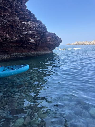 The image shows a close-up view of a teal paddleboard with black stripes floating on the surface of clear, calm water. The paddleboard has the text "WONDER IS ALL-AROUND" printed on its side. In the background, a large, reddish-brown cliff juts out into the water on the left side of the frame. The water is so clear that the rocky seabed is visible beneath the surface. Further in the distance, several white buoys are lined up on the water. The sky is a clear, bright blue. The scene is likely captured in the coastal waters near Sveti Stefan, Montenegro, given the characteristic rocky shoreline and clear Adriatic Sea.