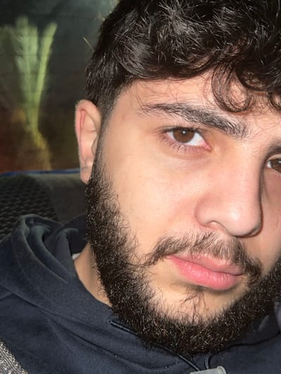 A young adult male is depicted in a close-up portrait, oriented forward and looking directly into the camera. He has fair to olive skin, dark brown curly hair, and a full, dark brown, curly beard and mustache. His eyebrows are thick and dark, and his eyes are dark brown; the sclera of his right eye shows minor redness. His nose is prominent, and his lips are full and closed. He is wearing a dark blue or black hooded sweatshirt. The background is out of focus, dark, and contains blurry, elongated, light-colored reflections with a greenish-yellow tint in the upper left, suggestive of foliage or an external light source viewed through a window. The environment provides no discernible specific identifying features to confirm a location such as Al-Muhajerin Municipality, Syria. The individual is static, posing for the image. No other subjects or interactions are present.