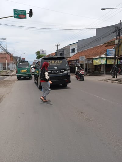 The scene takes place on a street in Katapang, Indonesia, located at coordinates -6.988 latitude and 107.558 longitude. A woman in a black hijab and red shirt is walking across the street. She is wearing blue jeans and sandals. A dark grey minivan with a white "MOUZA" logo and a license plate reading "D 1111 M.F" is parked on the right side of the street. A green van with a white license plate is parked on the left side of the street. A motorcycle is parked on the right side of the street. There is a green sign with white lettering that reads "FACTORY OUTLET" and an arrow pointing to the left, with a "Hilon" logo below it. There is a traffic light on the street. The street has a pothole in the middle of the road. There are power lines overhead. The scene is taken on a bright, sunny day.